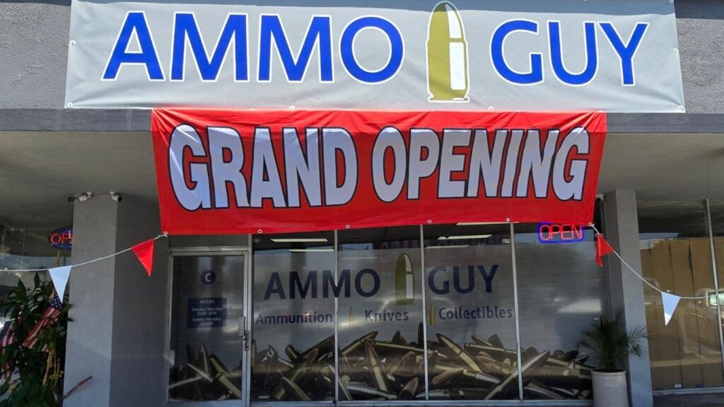 Front view of Ammo Guy's new ammunition store in Santa Ana, CA, featuring a large 'Grand Opening' banner in red and white. The store's sign, displaying 'AMMO GUY' with a bullet icon, is visible above the entrance, alongside a window showcasing 'Ammunition, Knives, Collectibles.'