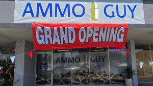 Front view of Ammo Guy's new Orange County ammo store in Santa Ana, CA, featuring a large 'Grand Opening' banner in red and white. The store's sign, displaying 'AMMO GUY' with a bullet icon, is visible above the entrance, alongside a window showcasing 'Ammunition, Knives, Collectibles.'
