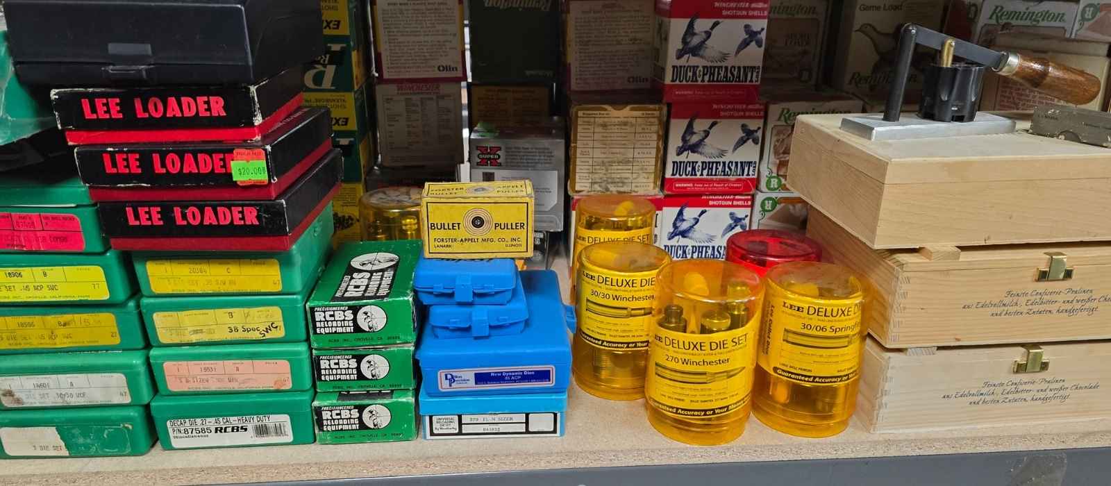 Interior view of ammo store featuring the reloading supplies area with various materials, equipment and tool kits for reloading ammunition lined up next to each other. A vintage RCBS reloading kits, a bullet puller and several die sets can be seen.